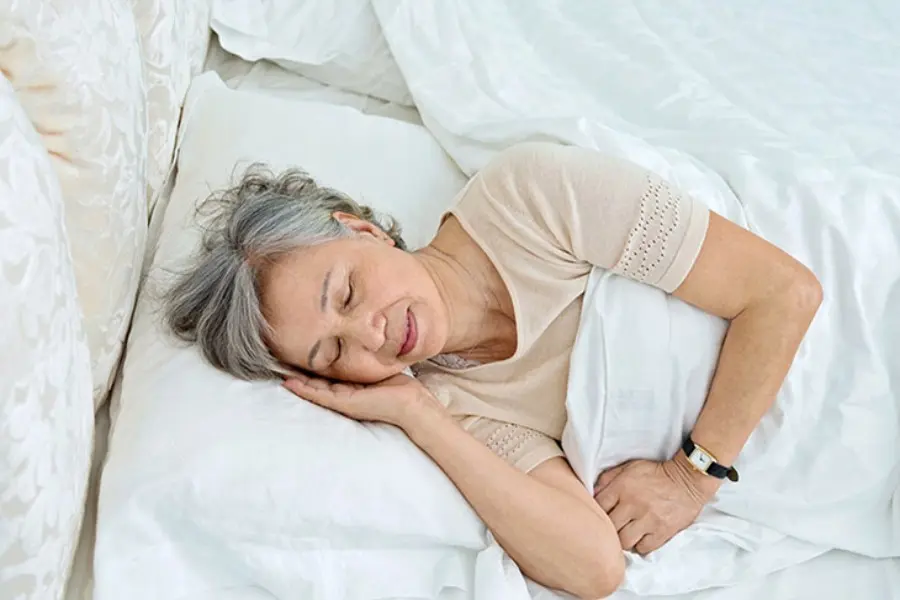 Cụ bà đang nằm ngủ trên giường với chăn và gối màu trắng ngà